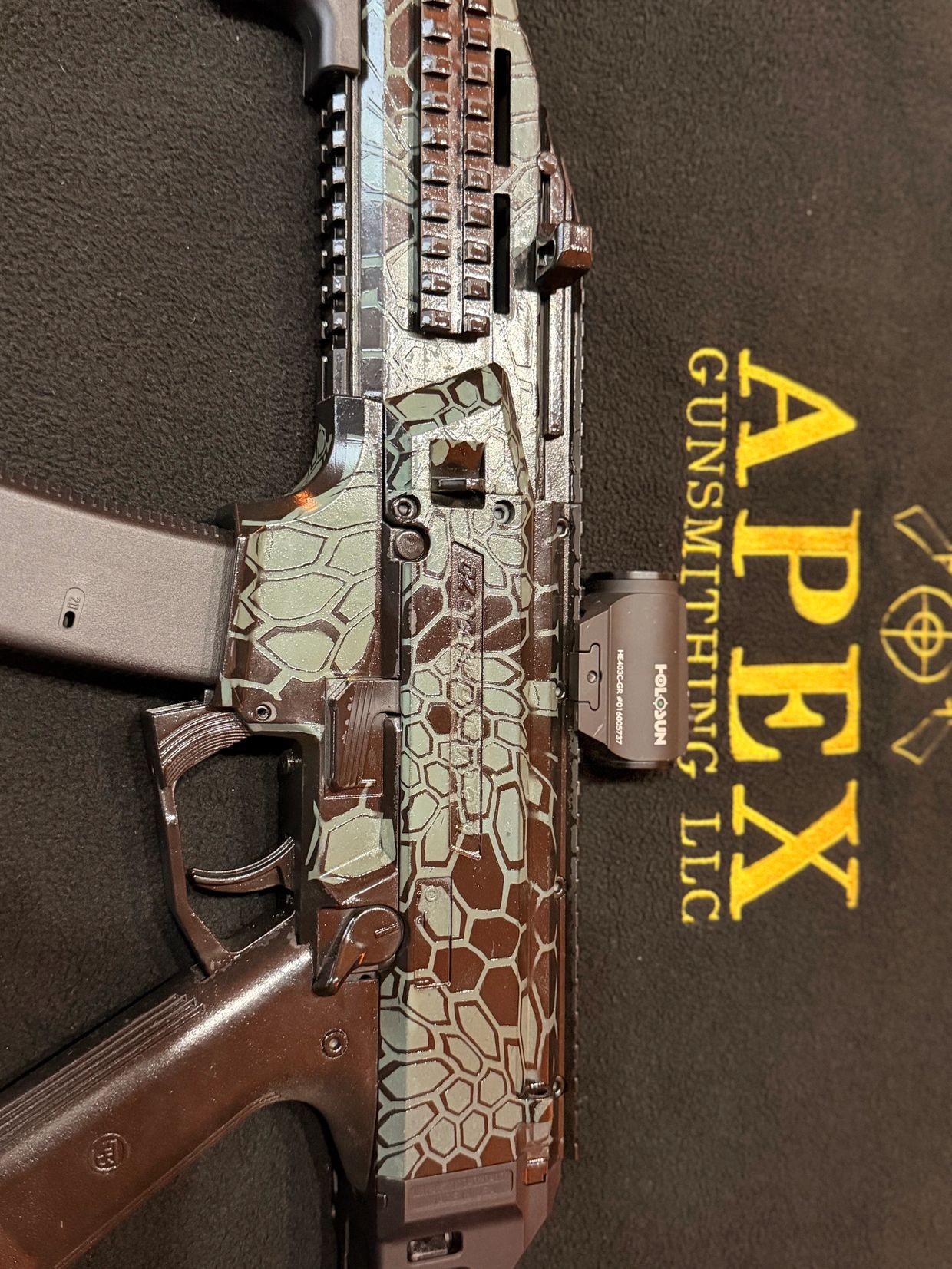 Close-up of a camouflaged firearm on an Apex Gunsmithing LLC mat.