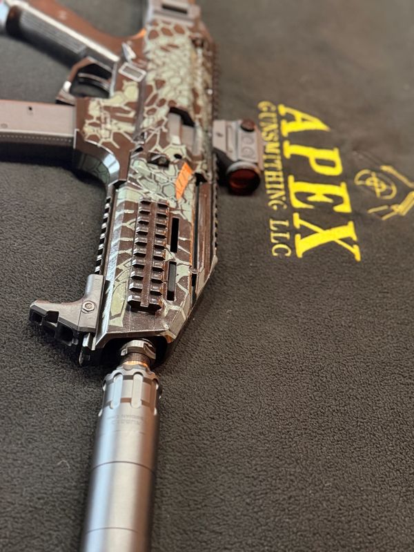 A camouflaged firearm with a suppressor on an Apex Gunsmith LLC mat.