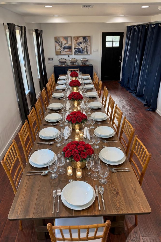 Elegant long dining table set with white plates, red rose centerpieces, and candles in a modern room.