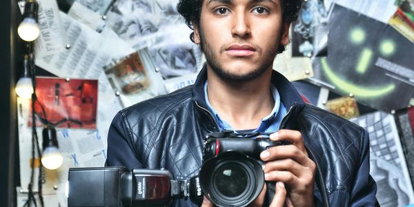 Young man holding a camera with decorative lights and pinned papers in the background.