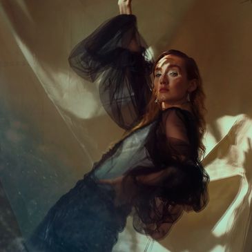 Woman in a sheer black dress poses gracefully in soft, moody lighting.