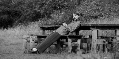 A woman leans backward on a wooden picnic table in a grassy area.