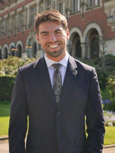 Smiling man in a suit standing outdoors in front of a historic building.