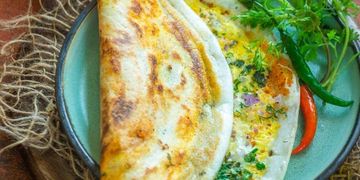 A delicious folded dosa filled with egg and herbs on a plate with green and red chilies.