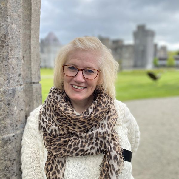Smiling woman in glasses and leopard print scarf outdoors near stone structure.