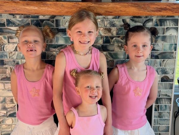 Four smiling girls in matching pink tops and white skirts with playful hairstyles.