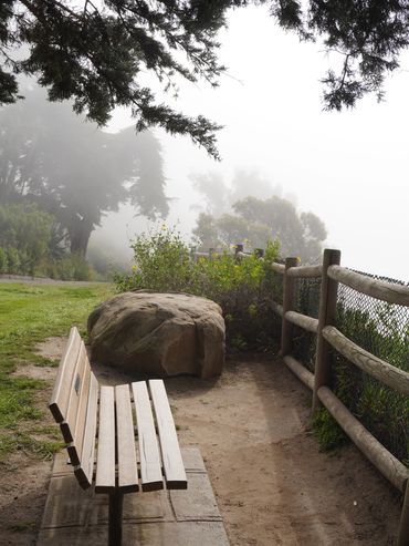 Empty park bench beside a dirt path and wooden fence on a foggy day.