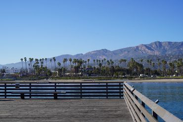Wooden pier overlooking a calm bay with palm trees and mountains in the background.