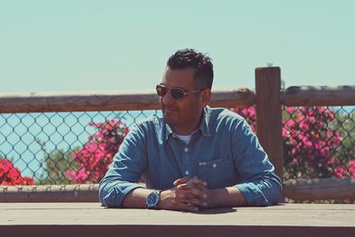 Man in sunglasses sitting at a wooden table outdoors with flowers in the background.