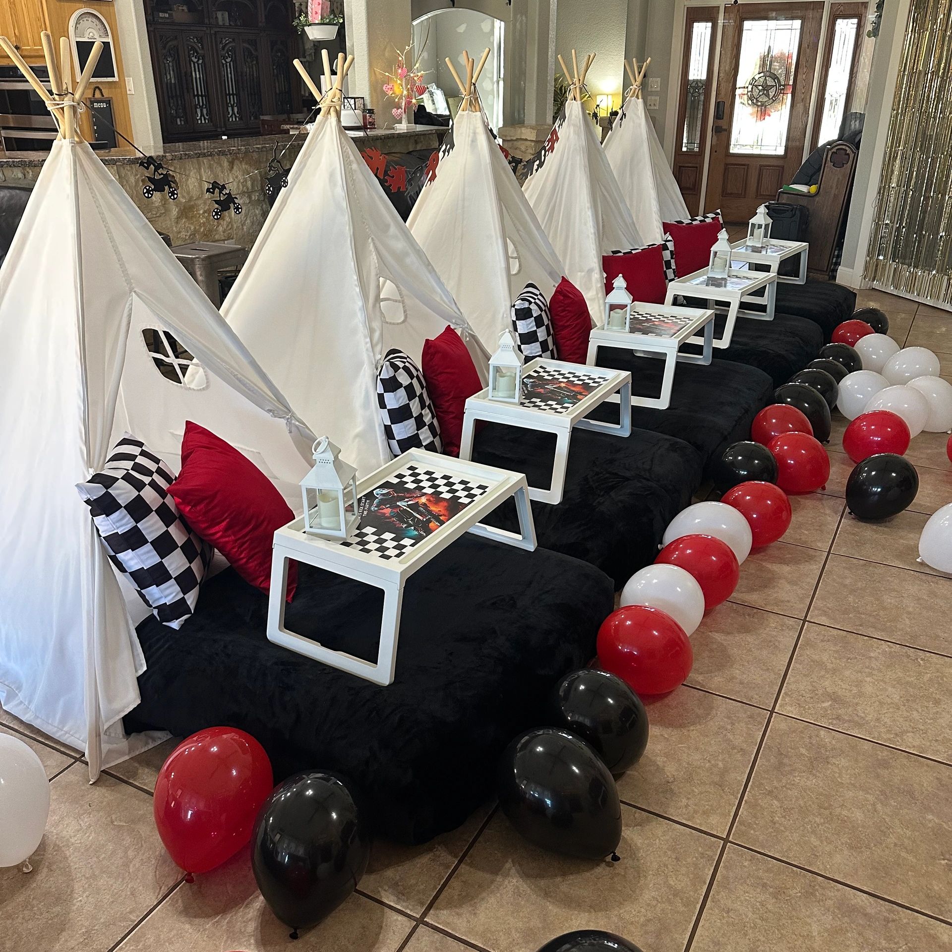 Indoor kids' party setup with white teepees, black cushions, and colorful balloons.