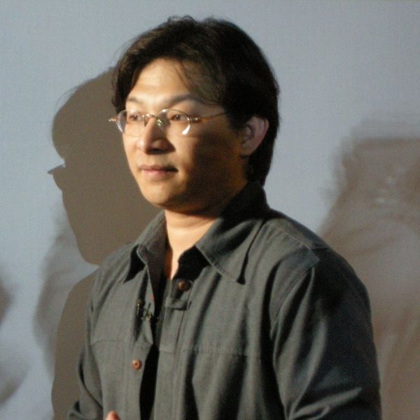 Man wearing glasses and a dark jacket stands against a plain background.