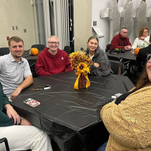Group of people sitting around a table with autumn-themed centerpiece and pumpkins in the background.