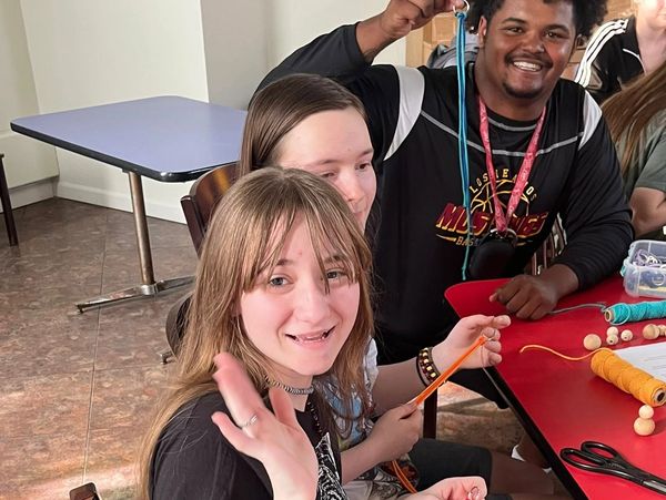 Three friends crafting with yarn and beads around a red table.