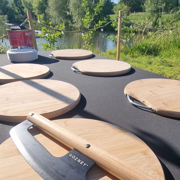 Wooden cutting boards and a mezzaluna knife on a table outdoors by a lake.