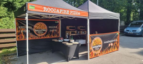 Outdoor pizza stall named ROCCAFIRE PIZZA with ovens and prep table.