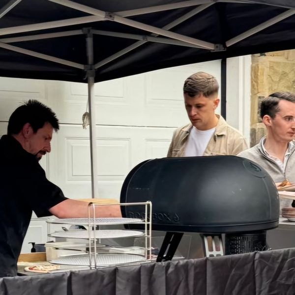 Three men working at an outdoor pizza oven setup under a tent.