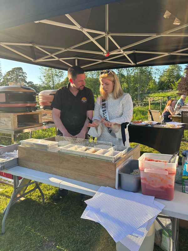 Two people discussing food toppings at an outdoor stall under a canopy.