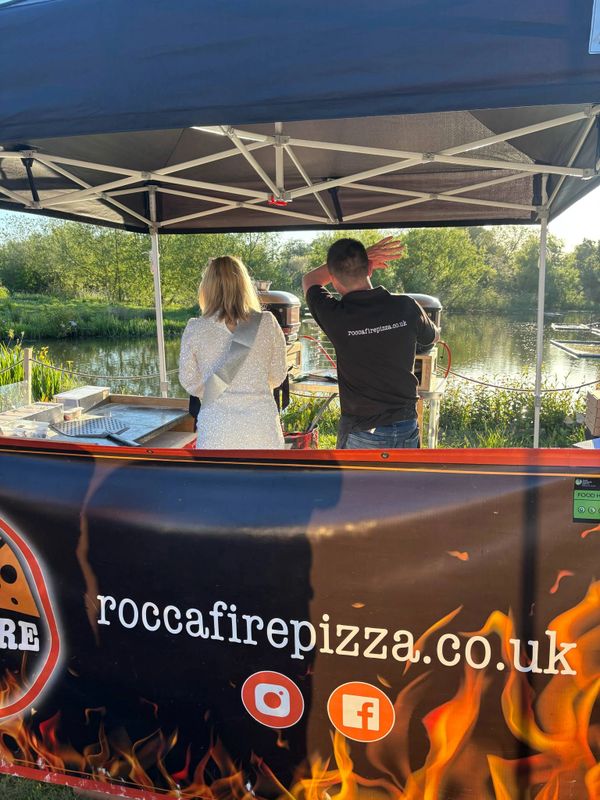 Two people working at a pizza stall by a lakeside under a canopy.
