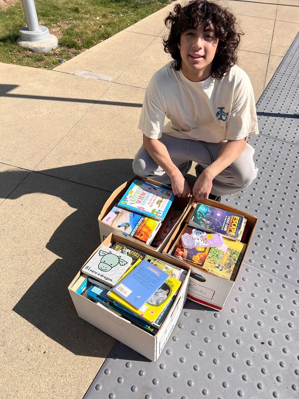 Young person kneeling beside three boxes filled with various books outdoors in sunlight.