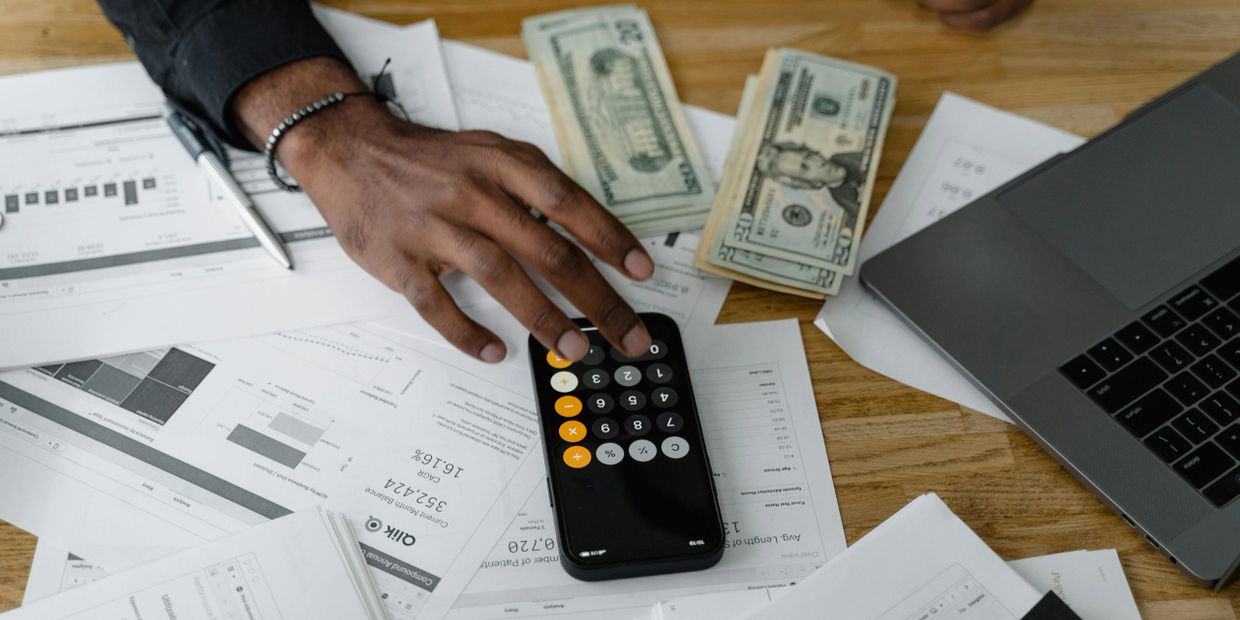 Person using a smartphone calculator amid documents, cash, and a laptop on a wooden table.