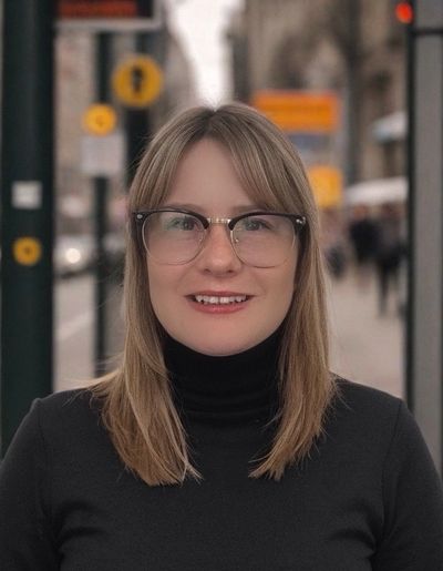 Smiling woman with glasses wearing a black turtleneck outdoors.