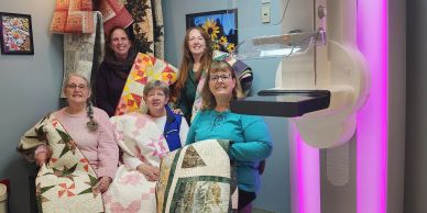 Five women proudly display their handmade quilts in a cozy room.