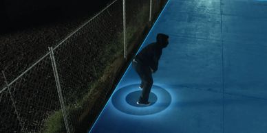 A person crouching on a blue surface with a glowing circle beneath them at night.