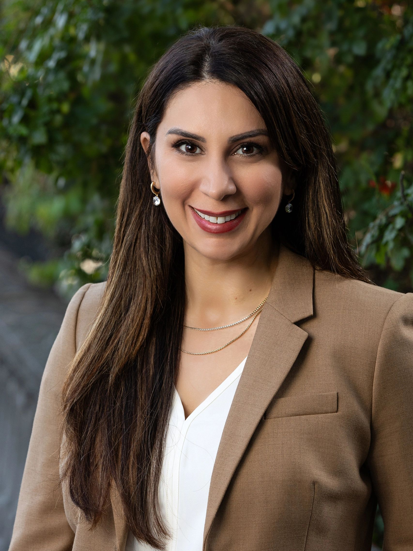 Professional woman in a brown blazer smiling outdoors with greenery background.