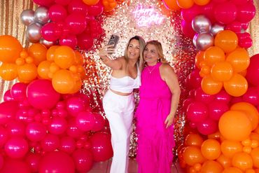 Two women posing for a selfie amidst bright balloon decorations.