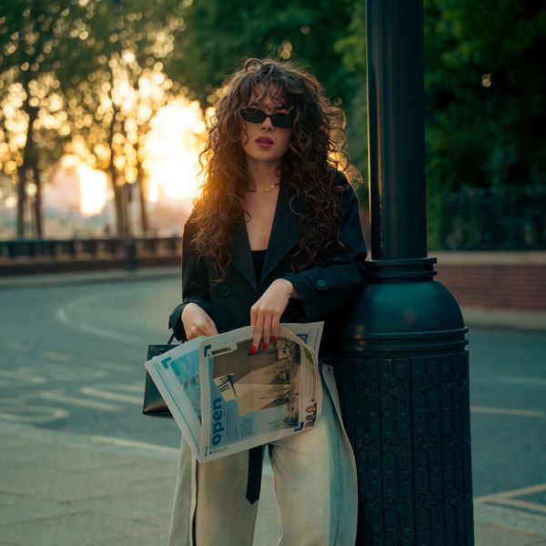 Woman in black coat holding newspapers by a streetlamp at sunset.