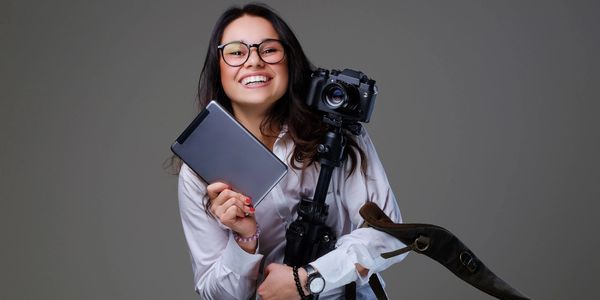 Smiling woman holding a tablet and a camera on a tripod, ready for creative work.