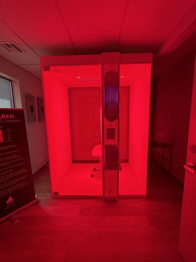 A red-lit therapy chamber with a chair inside and control panel outside.