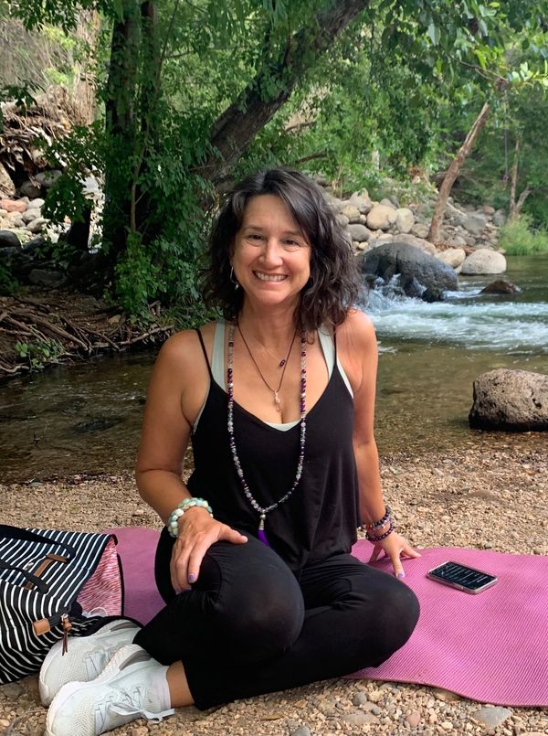 Woman sitting on a pink mat by a river, smiling at the camera.
