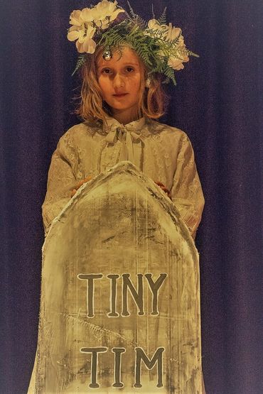 Girl wearing floral headpiece behind a tombstone labeled 'Tiny Tim'.
