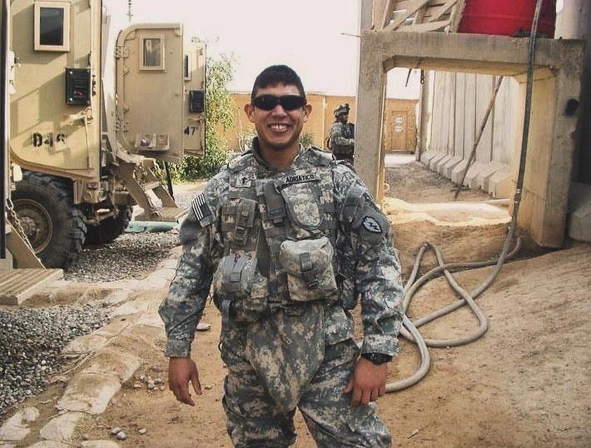 A smiling soldier in camouflage uniform and sunglasses outdoors.
