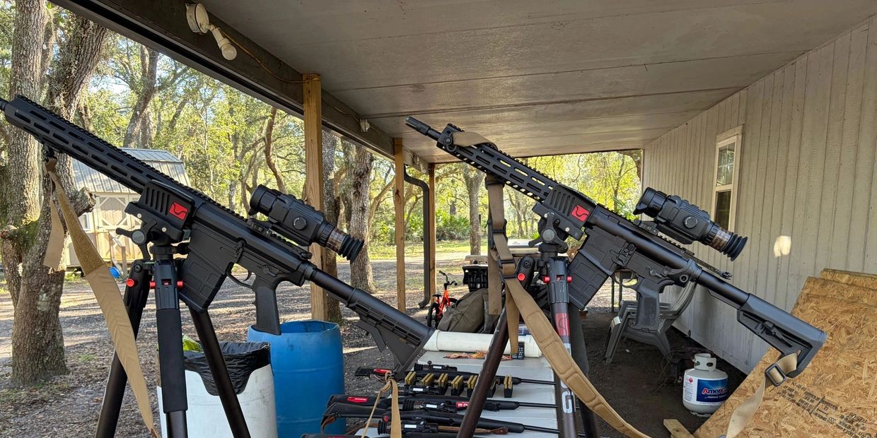 Two rifles set up on tripods under a patio with gear and supplies.