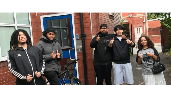 Five diverse friends posing outside a brick building with thumbs up and casual attire.