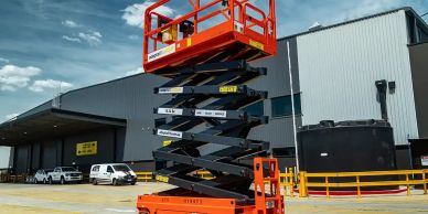 Orange scissor lift extended outside an industrial building under a partly cloudy sky.