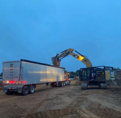 Excavator loading dirt into a truck at dusk on a construction site.