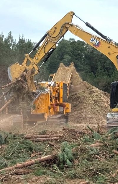 Excavator loading wood into a chipper, producing a large pile of wood chips.