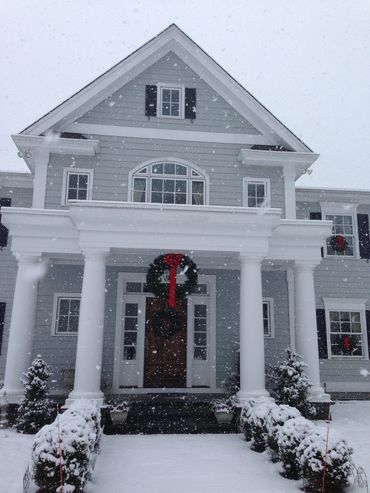 Snowfall on a decorated house with a Christmas wreath and snow-covered bushes.