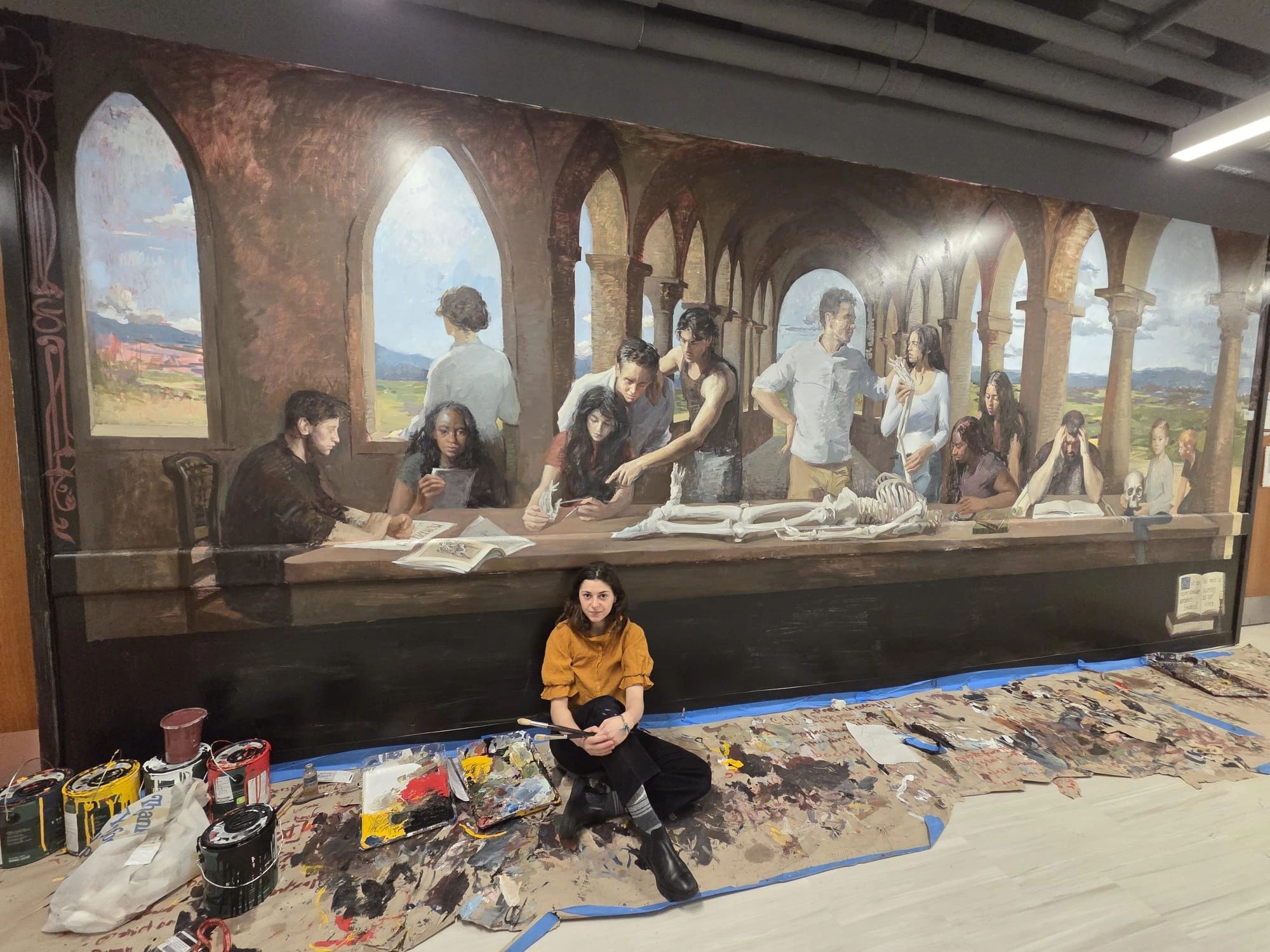 Artist sitting in front of a large mural with paint supplies.