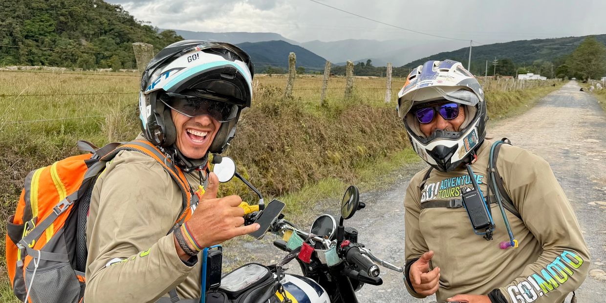 Two smiling motorcyclists in helmets on a rural road with mountains in the background.