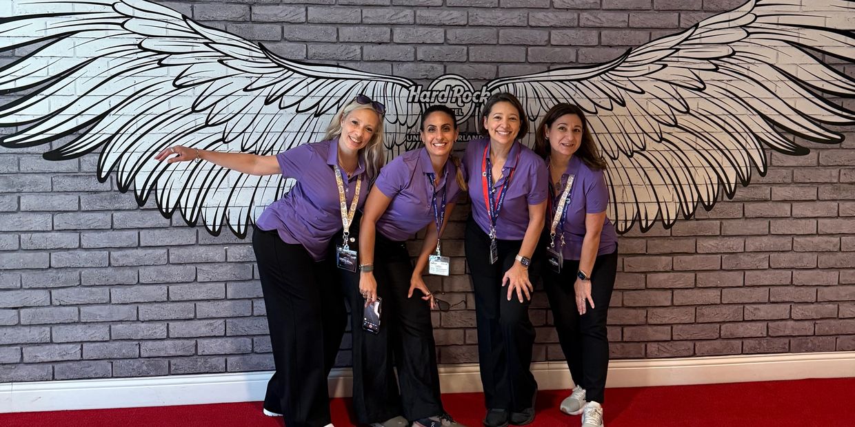 Four women in purple shirts posing with angel wings mural at Hard Rock Hotel.