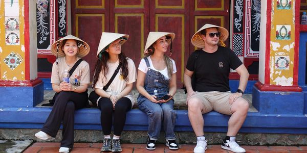 Four tourists wearing conical hats sit on steps in front of a colorful building.