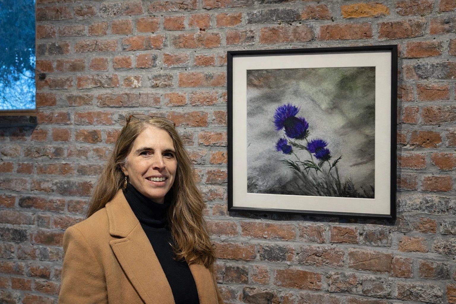 Woman standing beside framed purple flower artwork on brick wall.
