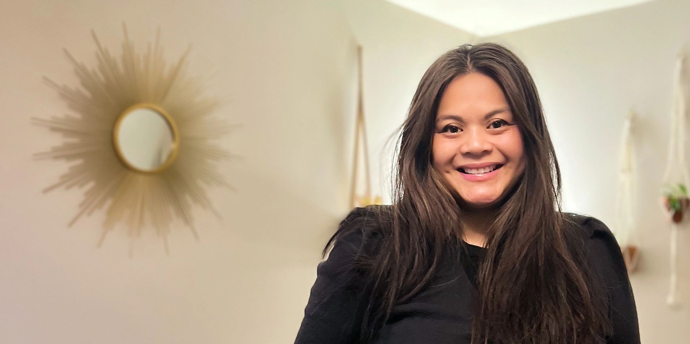 Eileen Espiritu, L.Ac. smiling. Standing in front of a cream wall, sun mirror and plants in the back