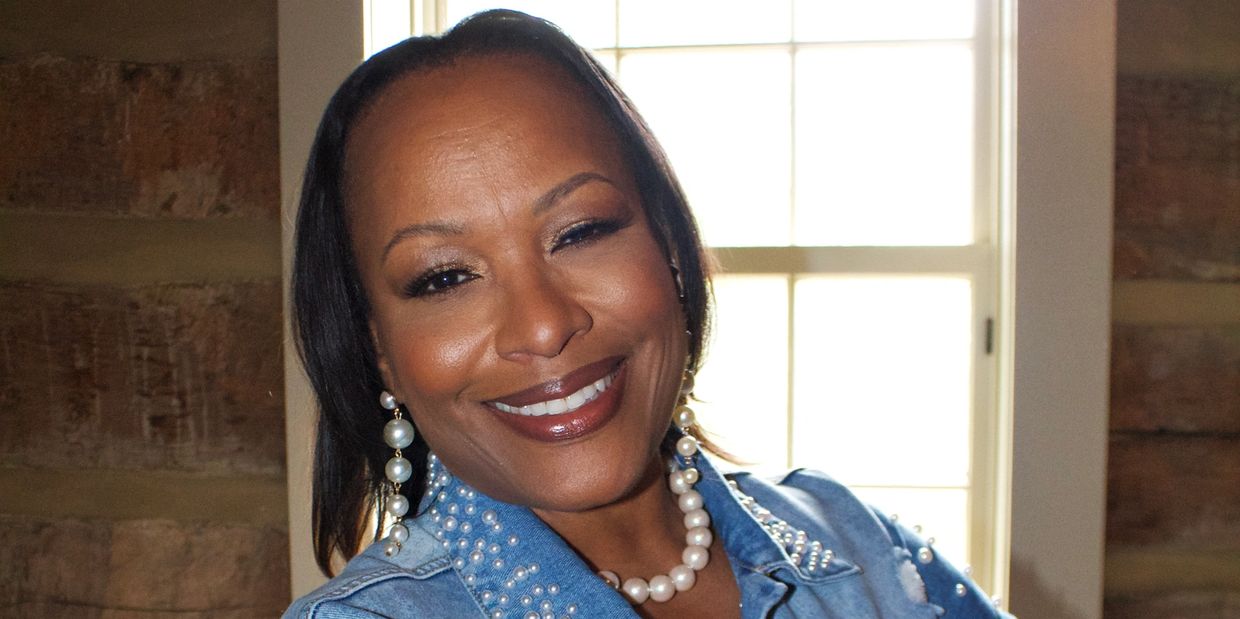 Smiling woman in pearl jewelry and denim jacket in front of a window.