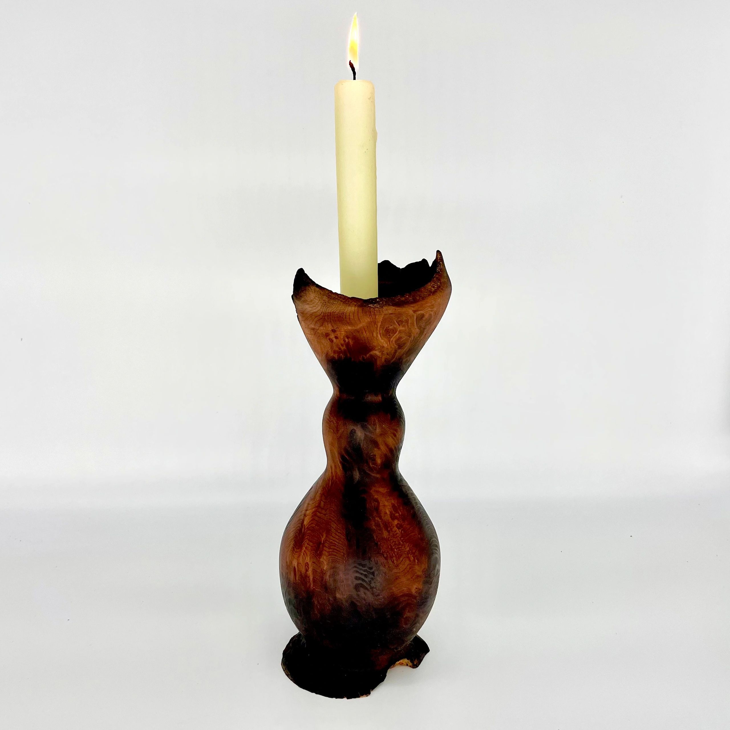 A wood turned candlestick holder made of redwood, holding a single long white candle that is lit.