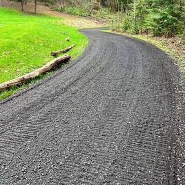 A freshly paved black gravel path curves through a green grassy area and forest.
